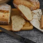 A wooden cutting board with a loaf of bread and two slices of bread, along with a bread knife. A proofing basket covered with a cloth and a bread lame are placed nearby on a dark rustic table, showcasing the beauty of homemade recipes.