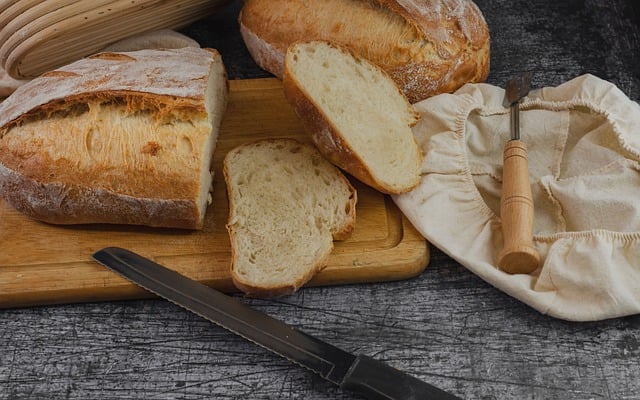 A wooden cutting board with a loaf of bread and two slices of bread, along with a bread knife. A proofing basket covered with a cloth and a bread lame are placed nearby on a dark rustic table, showcasing the beauty of homemade recipes.
