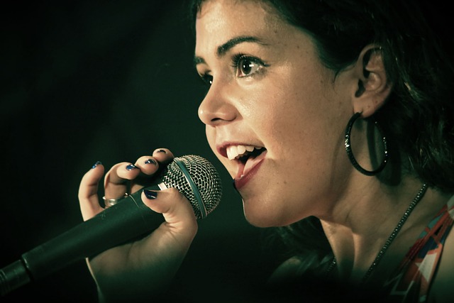 A woman with dark hair sings into a microphone, embodying the idea that anyone can learn singing. She has hoop earrings and painted nails, her face illuminated by the lighting as she appears enthusiastic and engaged. The dark background highlights her as the focal point.