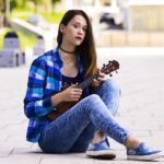 A young person with long hair, wearing a blue plaid shirt, jeans, and slip-on shoes, sits cross-legged on a paved path, playing a ukulele. They are outdoors, with buildings and greenery in the background—an ideal scene for beginners to learn ukulele.