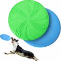 A black and white dog leaps joyfully with its mouth open, aiming to catch a light blue frisbee. Behind the dog, there are two stacked frisbees, one green on top and one blue underneath, both with a textured design pattern. The background is white, capturing the essence of joyful frisbee playtime.